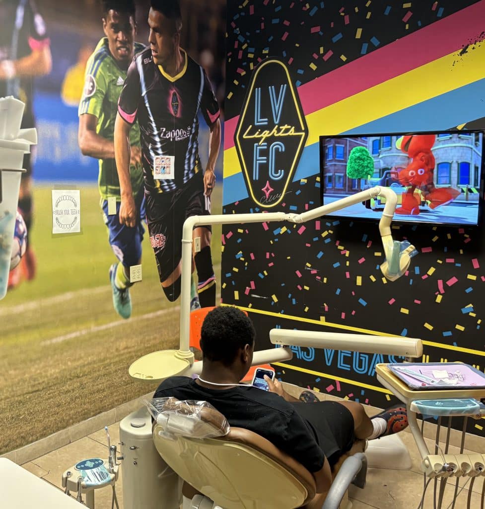 Affordable Dental for Kids soccer room, featuring a brightly colored dental office room with a pro soccer image on the wall. A young patient sits in the chair, waiting for the dentist, checking his phone while watching something on the TV