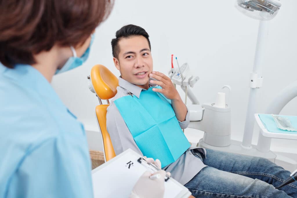 Asian male patient sitting in a dentist office chair lightly touches his jaw, explaining dental pain to a dentist in view, prescribing medication for an infected cavity