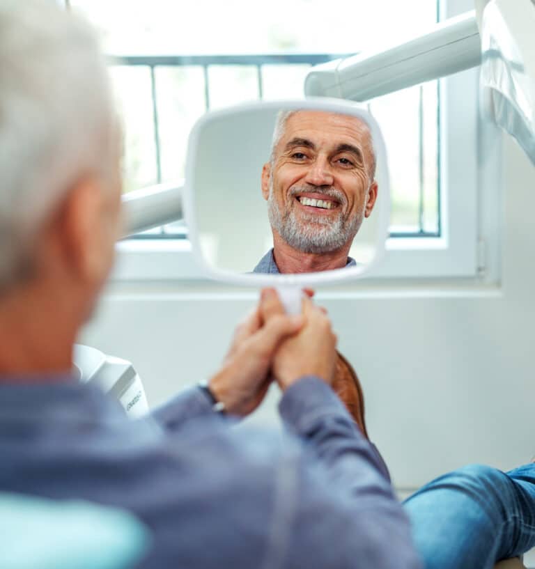 Handsome middle aged man smiles at his face from a square shaped mirror in a dentist office room, displaying straight white teeth