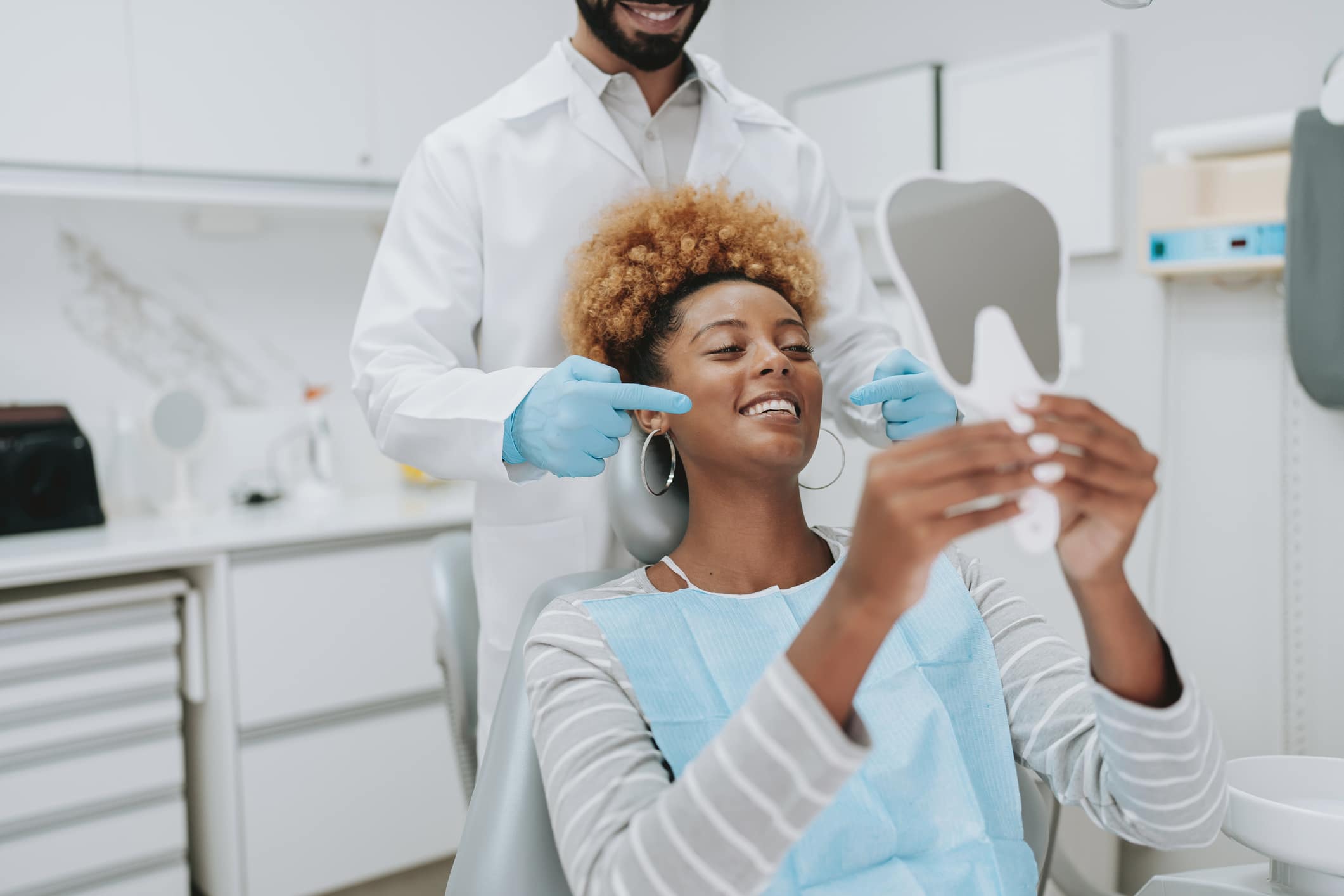 Beautiful dark skinned woman with large hoop earrings smiling with bright, white teeth and looking into a tooth-shaped mirror in a dentist examination room, a bearded male dentist behind her, pointing to her teeth with light blue gloves